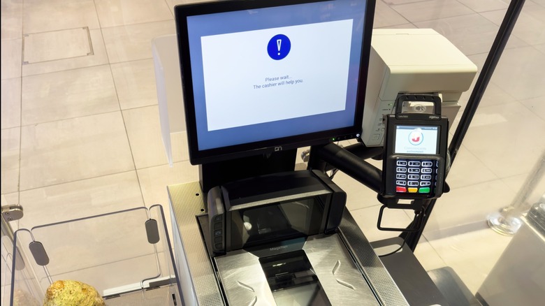 A self-checkout machine in a French supermarket shows a message awaiting cashier help, with scanned items including vegetables and packaged food visible
