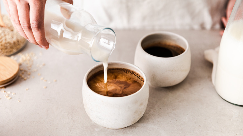 A hand is pouring a bottle of milk into a cup of black coffee in a white cup