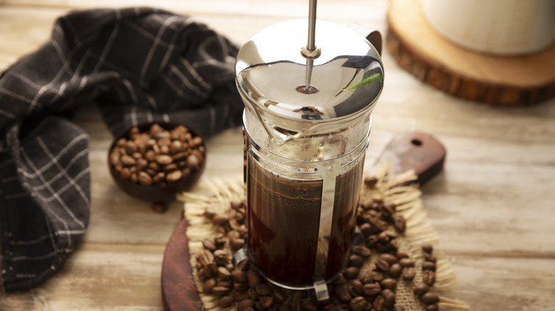 A French press coffee maker on a wooden table, surrounded by coffee beans and a dishtowel
