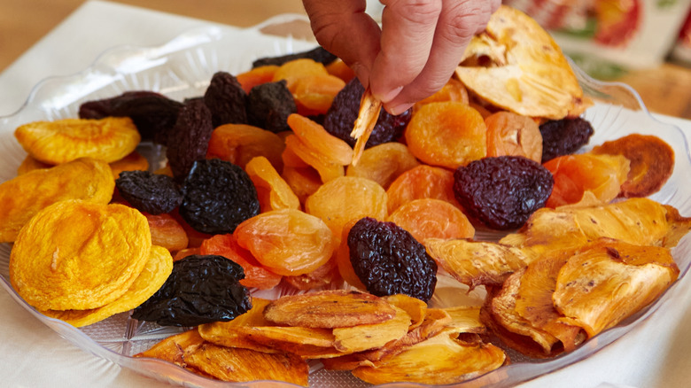 Hand selecting various dried fruits from a clear plate, including apricots, prunes, and apple slices, arranged on a white tablecloth