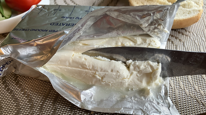 a knife cuts into a brick of bettergoods cream cheese oozing whey onto the foil wrapper