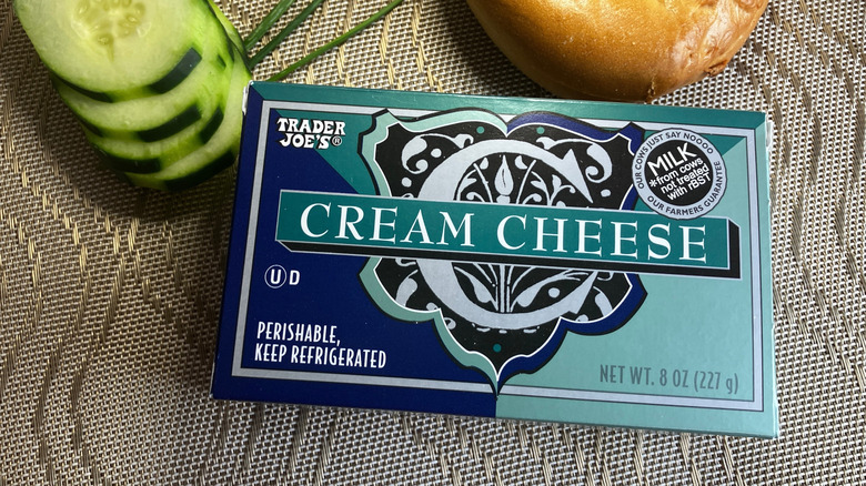 close up of blue and green box of Trader Joe's cream cheese with sliced cucumbers and a bagel in the background