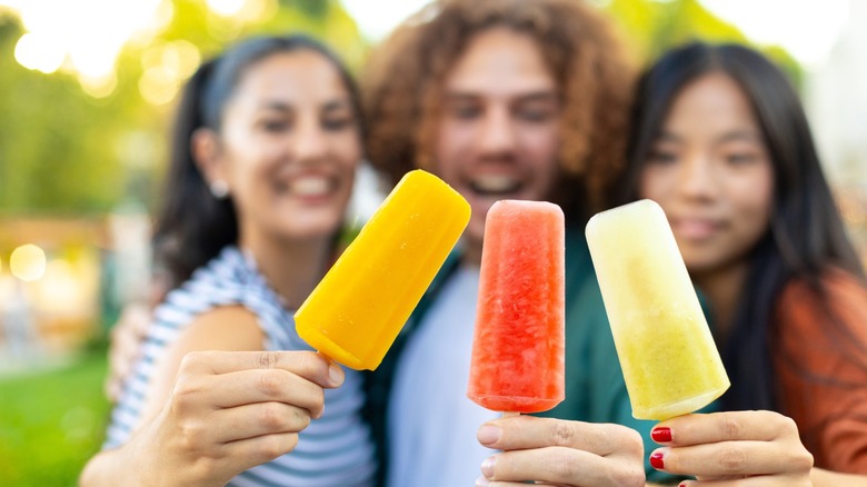 Three people holding colorful ice cream popsicles on a sunny day outdoors