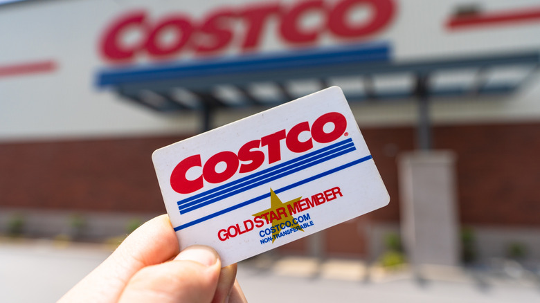 A hand holds a Costco card in the air, the blurred out storefront of Costco behind it.