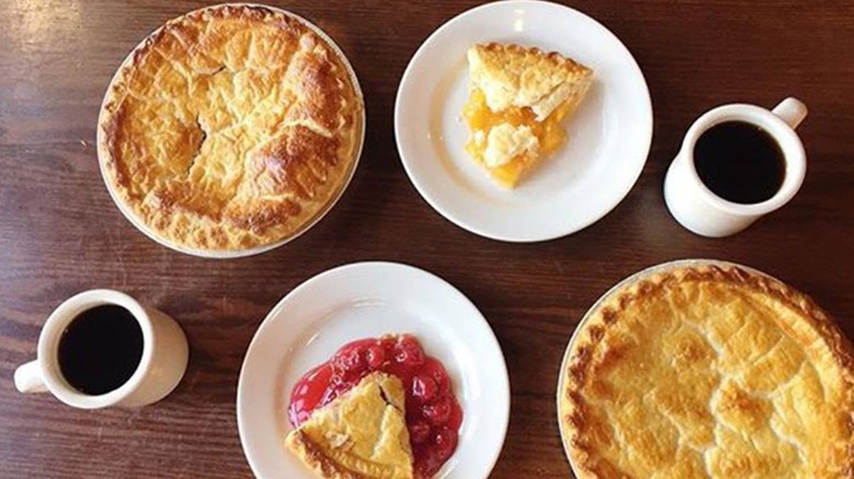 Marie Callender's assorted whole pies and pie slices on plates beside coffee mugs on wooden table at a Marie Callender's