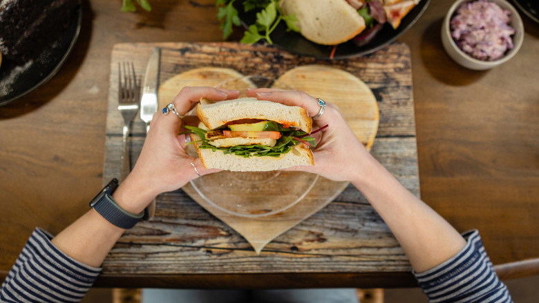 First-person point of view of a sandwich in a woman's hands, white bread with lettuce and tomatoes, over a heart-shaped plate