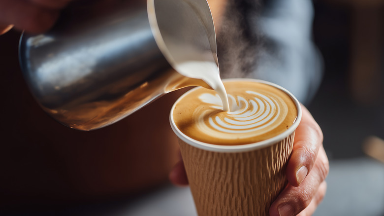 A barista pouring milk out of frothing pitcher to create design on surface of latte in takeout cup