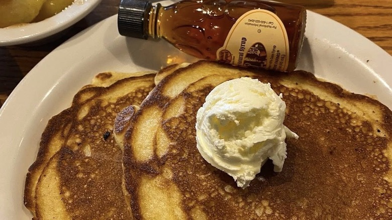 Two pancakes with a dollop of butter on top and a small jar of maple syrup on a white platter at Cracker Barrel.