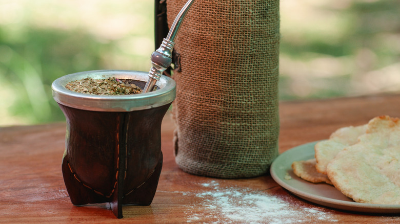 A cup of yerba mate with a bombilla inside rests on a wooden table. A bottle is behind it, and a plate of cake to the right.