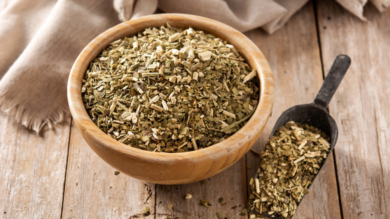 A wooden bowl of yerba mate leaves on a wooden table. A metal scoop filled with the product sits to the right.
