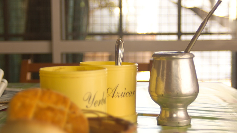 A cup of yerba mate with a bombilla. To the left is a tub of sugar with a spoon in it, and a pastry.