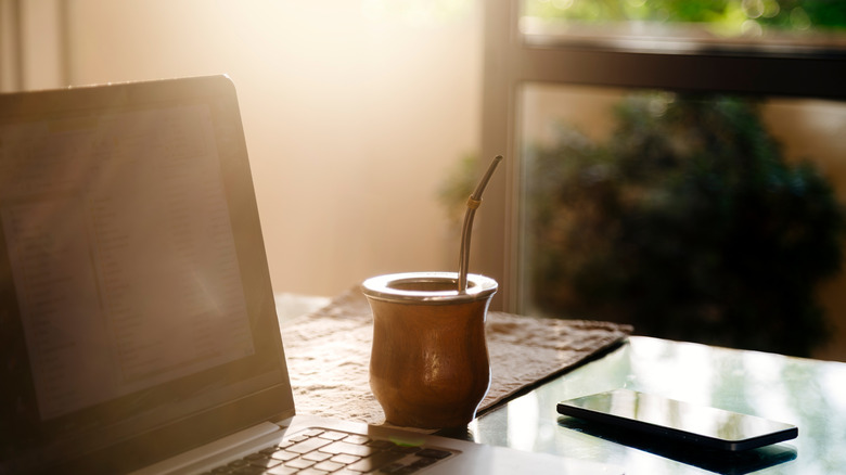 A traditional glass of yerba mate rests on a table with a straw in it. A cellphone and laptop surround the glass.