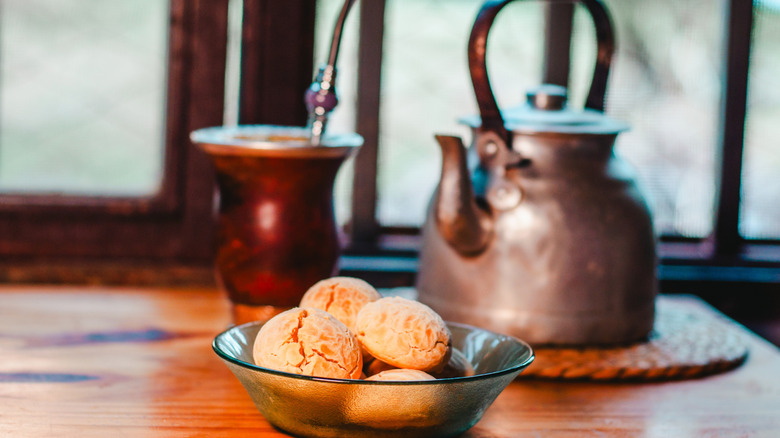 A cup of yerba mate sits beside a metal kettle on a trivet. A bowl of pastries sits in front of both.