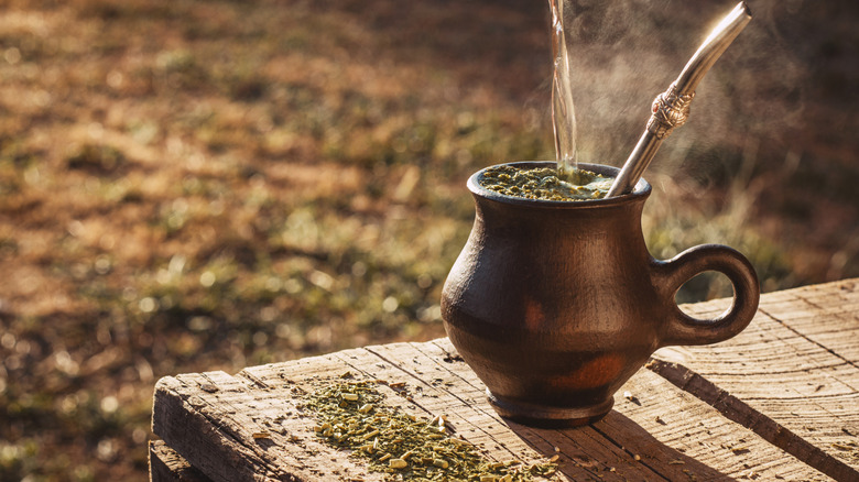 A cup of yerba mate rests on a wooden table with a bombilla inside. Water is being poured in the cup.