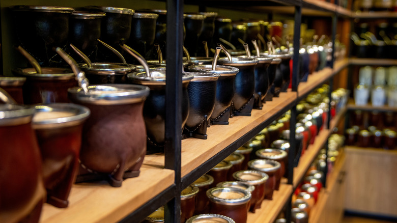 Several rows of mate gourds are lined up on wooden shelves, with bombillas inside of them.