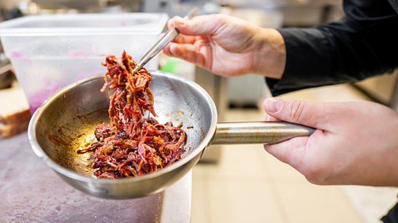 A professional chef holds a stainless steel pan filled with seasoned, shredded meat using tongs in a commercial kitchen