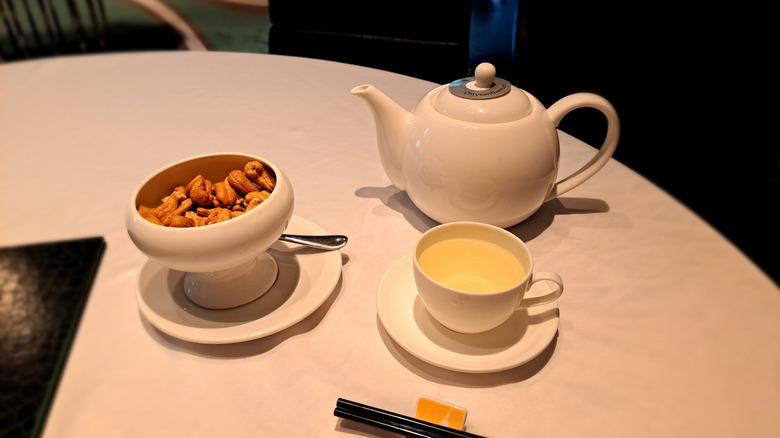 A teapot, teacup filled with mild tea, and a bowl of nuts on a white table
