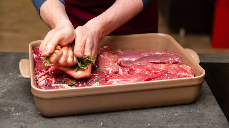 Hands squeezing rosemary and sage over raw beef cuts in a baking dish