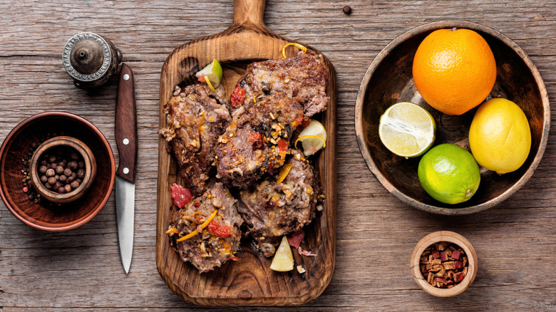 Cooked beef steaks on a wooden board next to bowls of citrus fruits and seasonings