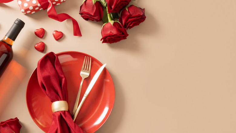 Valentine's day table setting for date with red plate, chocolate sweets, wine, romantic gift and red roses on beige background.