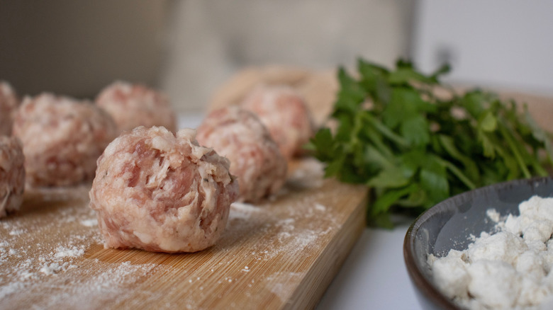 Raw meatballs on a wooden cutting board surrounded by parsley and cheese