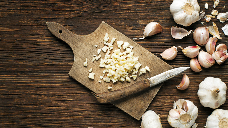 Sliced garlic on a wooden cutting board surrounded by garlic cloves and whole heads of garlic
