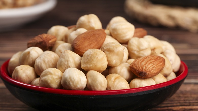 Roasted almonds and hazelnuts in a red and black bowl on wooden surface with blurred snacking ingredients in background