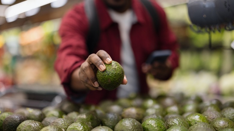 Hand reaching for a fresh avocado from a large display in the produce section of a grocery store