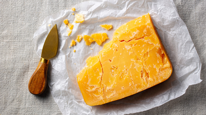 A slice of aged cheddar cheese on a square of parchment paper. A cheese knife rests to the left side.