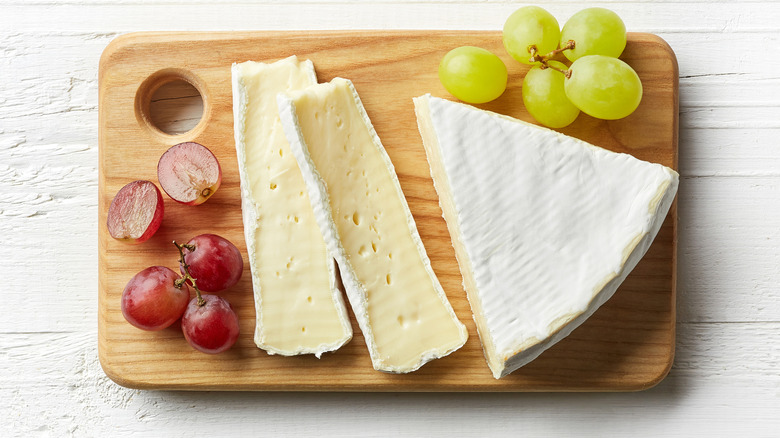 Pieces of brie sit on a wooden cutting board surrounded by grapes on either side