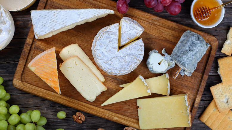 A spread of cheeses on a cutting board, surrounded by fruit, crackers, and a bowl of honey