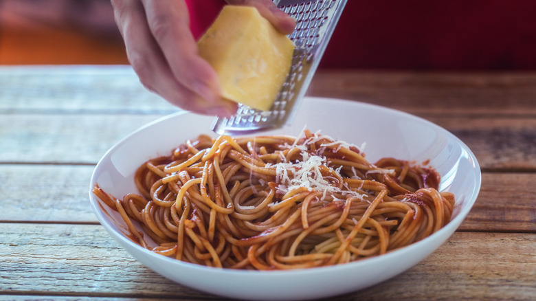 A hand grates a block of Parmesan onto a bowl of spaghetti