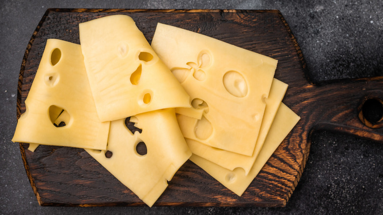 Several slices of Swiss cheese rest on a wooden cutting board