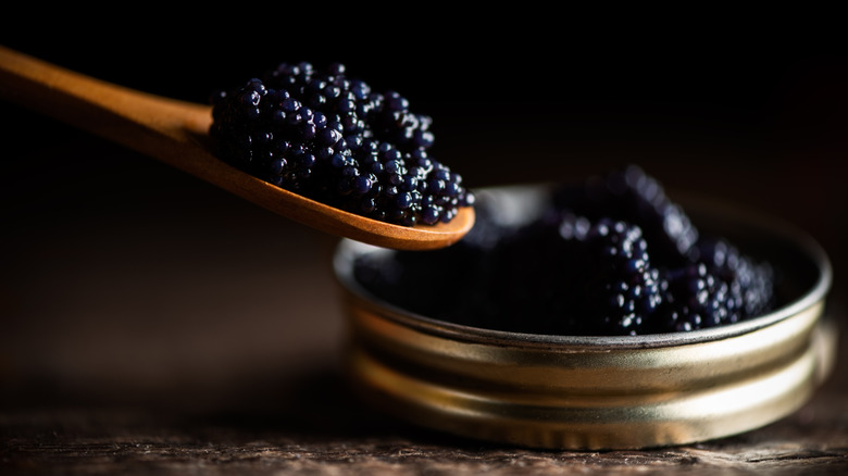 A tin of lumpfish roe sits on a table. A wooden spoon to the left is piled high with the roe.