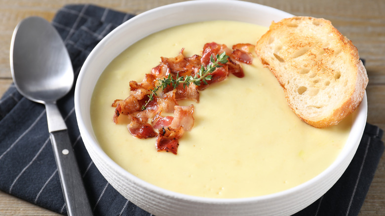 Potato soup in white bowl topped with bacon and a slice of bread