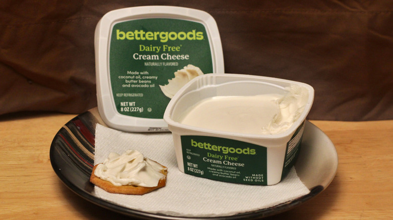 A container of Bettergoods Dairy Free cream cheese on a plate beside a cracker spread with the cream cheese