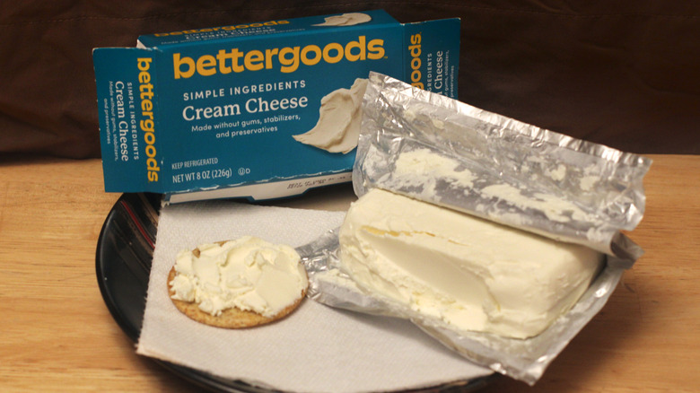 A brick of Bettergoods Plain Cream Cheese cheese on a plate beside a cracker spread with the cheese and the box it came in