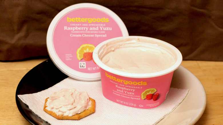 A container of Bettergoods Raspberry and Yuzu cream cheese spread on a plate beside a cracker spread with the cream cheese