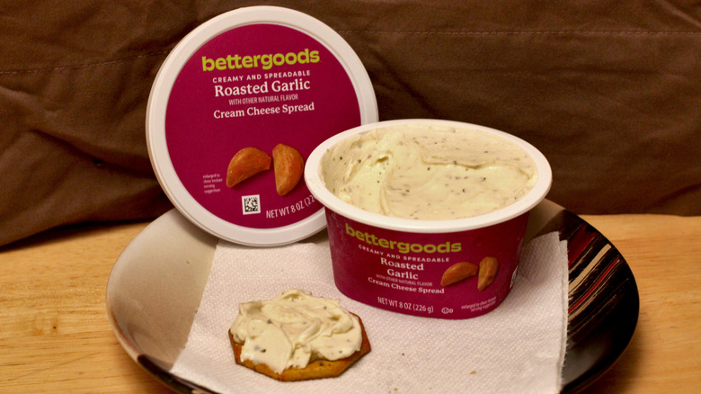 A container of Bettergoods Roasted Garlic cream cheese spread on a plate beside a cracker spread with the cream cheese