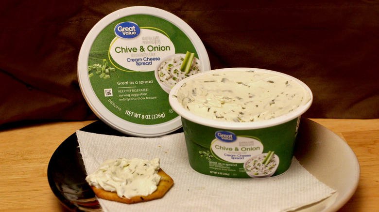 A container of Great Value Chive & Onion cream cheese spread on a plate beside a cracker spread with the cream cheese