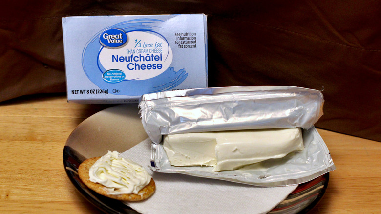 A brick of Great Value Neufchâtel cheese on a plate beside a cracker spread with the cheese and the box it came in