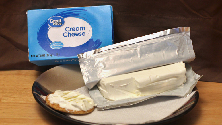 A brick of Great Value plain cream cheese on a plate beside a cracker spread with the cream cheese and the box it came in