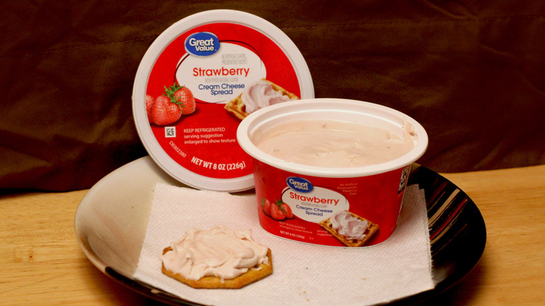 A container of Great Value Strawberry cream cheese spread on a plate beside a cracker spread with the cream cheese