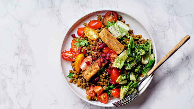 A grain bowl topped with colorful vegetables, including tomatoes, pomegranate seeds, and fried tofu