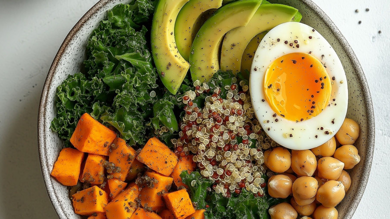 A grain bowl filled with various ingredients, including half a soft-boiled egg, chickpeas, roasted squash, kale, avocado, and quinoa