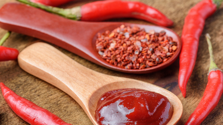 Wooden spoons with chili pepper flakes and paste on a wooden surface surrounded by whole, fresh chili peppers