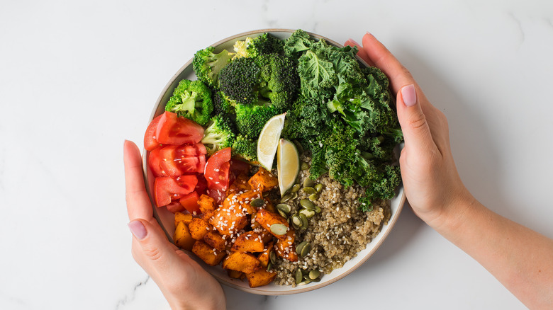 A woman's hands around a colorful grain bowl filled with broccoli, kale, tomatoes, sweet potatoes, pumpkin seeds, and lime wedges