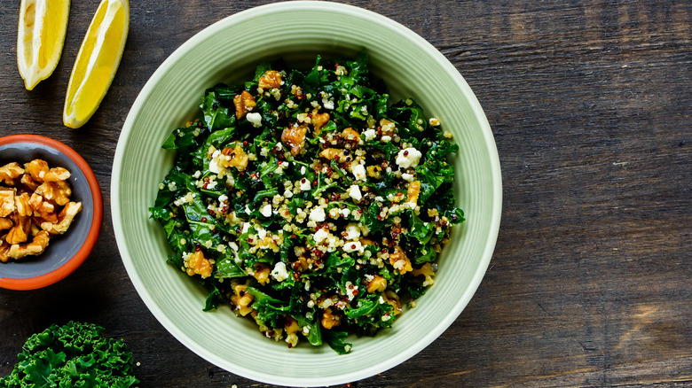 A grain bowl filled with green, leafy vegetables, topped with nuts on a wooden surface, garnished with lemon wedges