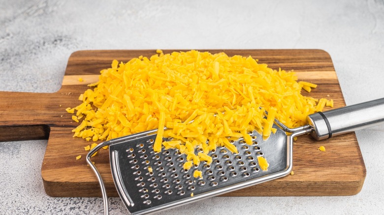 Cheddar cheese shreds and steel grater on wooden board over gray surface cooking ingredient texture for recipes and menus.