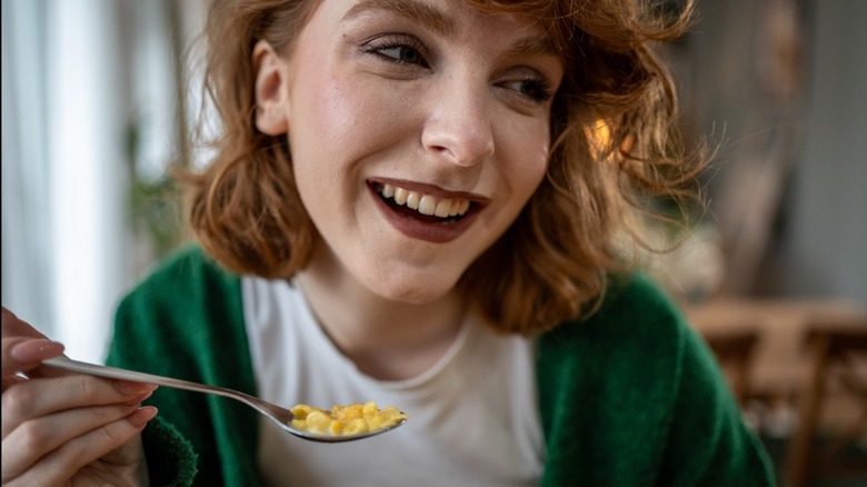 Young woman with vibrant red hair, wearing a cozy green cardigan, smiling joyfully while savoring a spoonful of creamy macaroni and cheese, enjoying a delightful meal at home